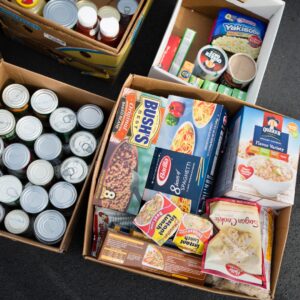 A bird's eye view shows open cardboard boxes filled with non-perishable food items, such as baked beans and oats.