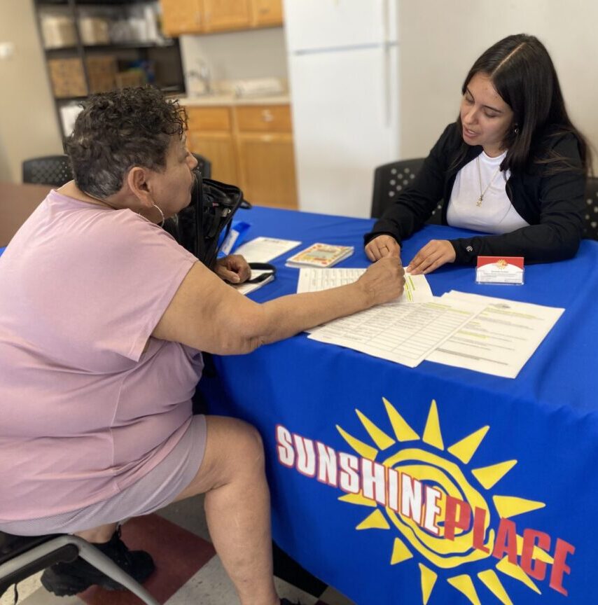 Two women sitting at a table with a blue tablecloth that reads "Sunshine Place" discussing housing options.