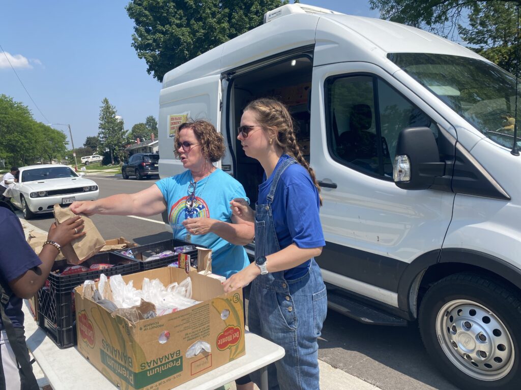 Two women hand out food in front of a white van with the Sun Prairie Food Pantry logo on the side.