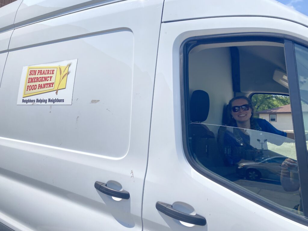 A smiling woman peers out the half-open window of a white van with a Sun Prairie Food Pantry logo on the side.