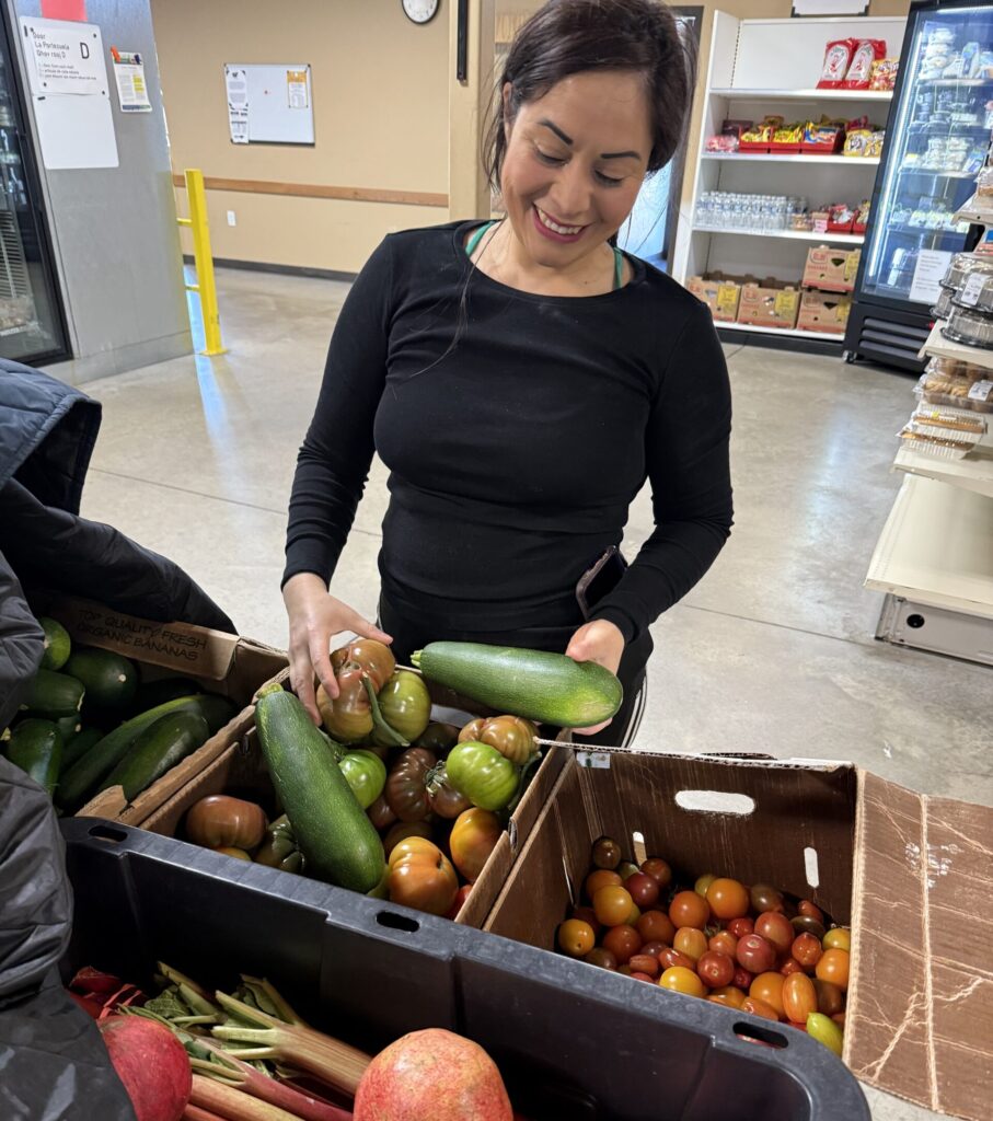 A smiling woman looks down at bins of fresh produce, including tomatoes, zucchini, and rhubarb.