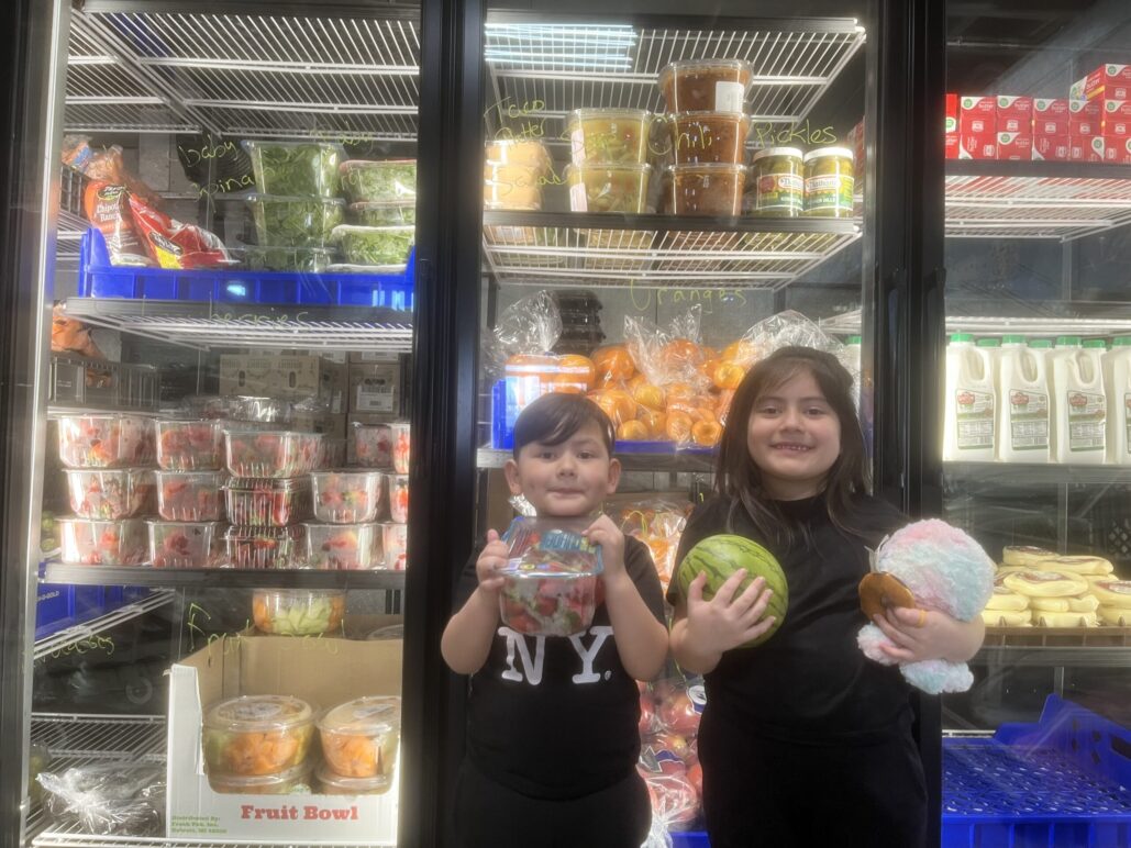 Two children stand side-by-side in front of a coolor of food holding fruit.