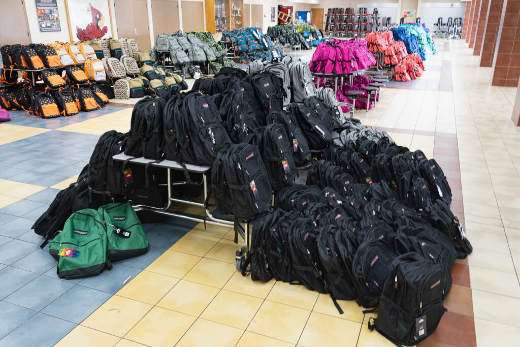 A long room that looks like a school cafeteria has backpacks of all colors sitting on the tables.