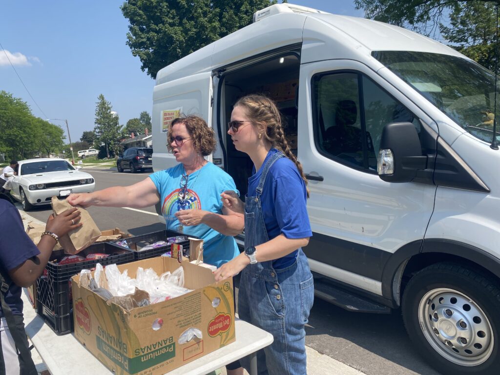 Two women stand by the white food pantry van and hand out food.
