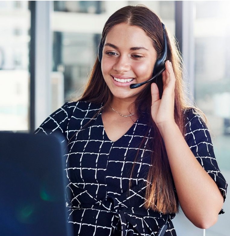 A smiling woman with long brown hair looks at a computer and speaks on a headset.