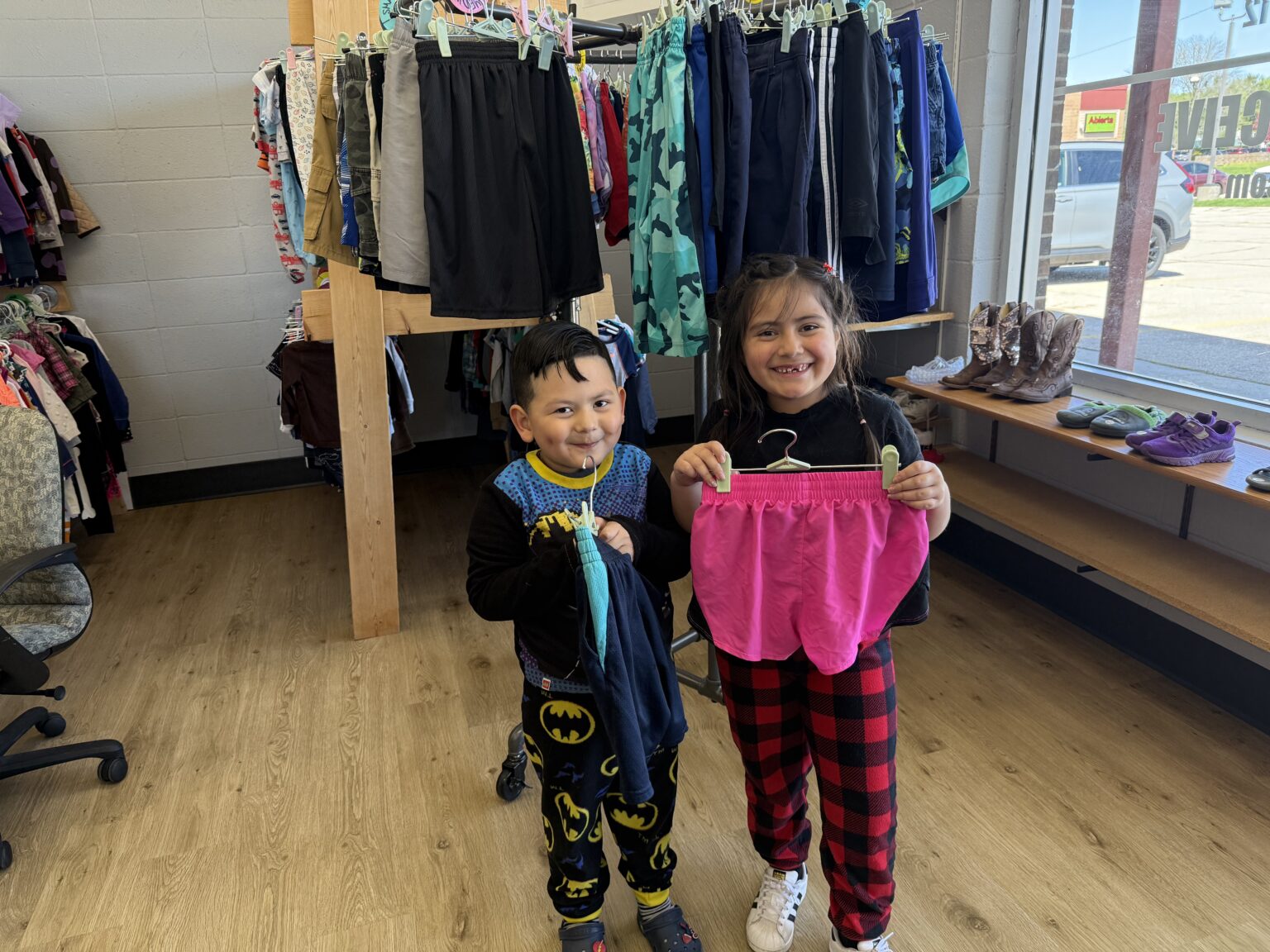 Two children smile and hold up clothing items inside of CARDS Closet.