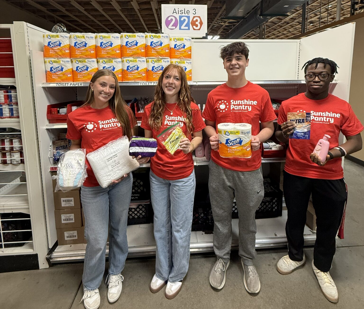 4 teens stand in front of a shelf of food in red Sunshine Pantry shirts, holding grocery items.