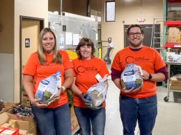 A group of 3 people wearing bright orange Bank of Sun Prairie shirts are holding packaged turkeys.