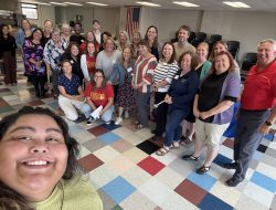 A smiling woman close to the lens takes a selfie with a large group of people behind her.