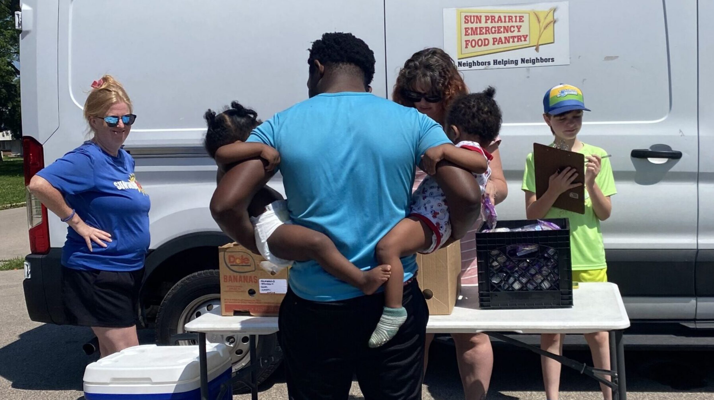 Summer Lunch 1 a father stands with a child on each hip in front of the food pantry truck and staff and volunteers handing out summer lunch bags.